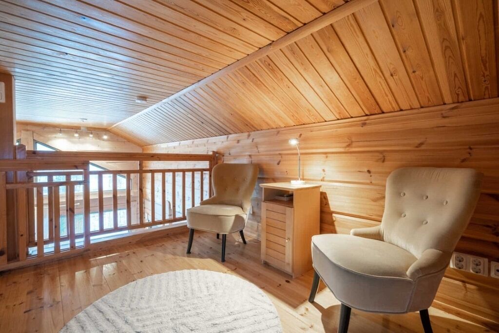 A cozy wooden cabin with a high ceiling, a balcony with a wooden railing, two upholstered chairs, and a small wooden cabinet.