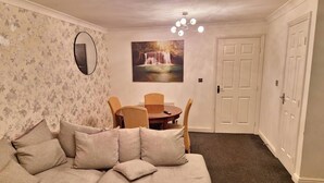 A cozy living room with a beige sofa, a dining table with chairs, a wall-mounted TV, and a painting of a waterfall. - 3 Bed House - Sleeps 6 - Parking - Wifi (Maidstone)