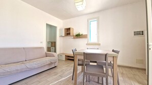 A modern, minimalistic living space with a sofa, a dining table, and chairs. There is a window providing natural light, and built-in shelving above the table.
