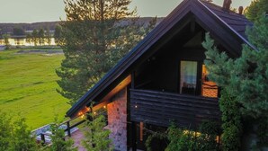 A modern cabin with a sloped roof, stone foundation, and a deck overlooking a grassy field.