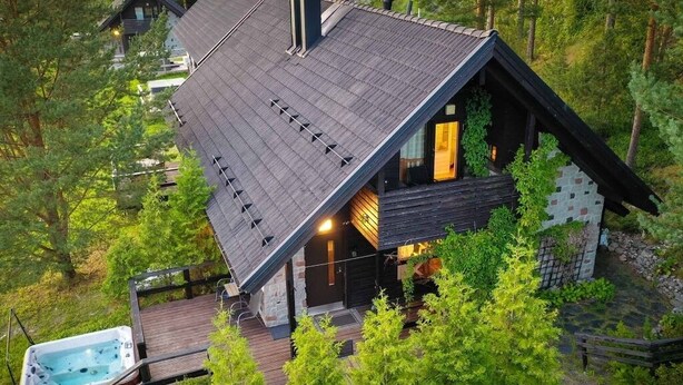 A modern cabin with a sloped roof, a hot tub on a wooden deck, and surrounded by dense forest.