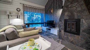 A cozy living room with a stone fireplace, a flat-screen TV, and a large window with a view of trees.