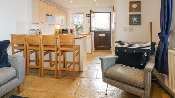 Ein gemütliches Wohnzimmer mit grauem Sofa, einem Holzesstisch mit Stühlen, einem kleinen Sessel mit blau-weiß gestreiftem Kissen und einer Küchenzeile mit Holzschränken und einem Fenster.