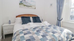 Ein Schlafzimmer mit karierter Bettdecke, einem Holzkopfende, einem Nachttisch mit Lampe und einem Bild eines Strandhauses an der Wand.
