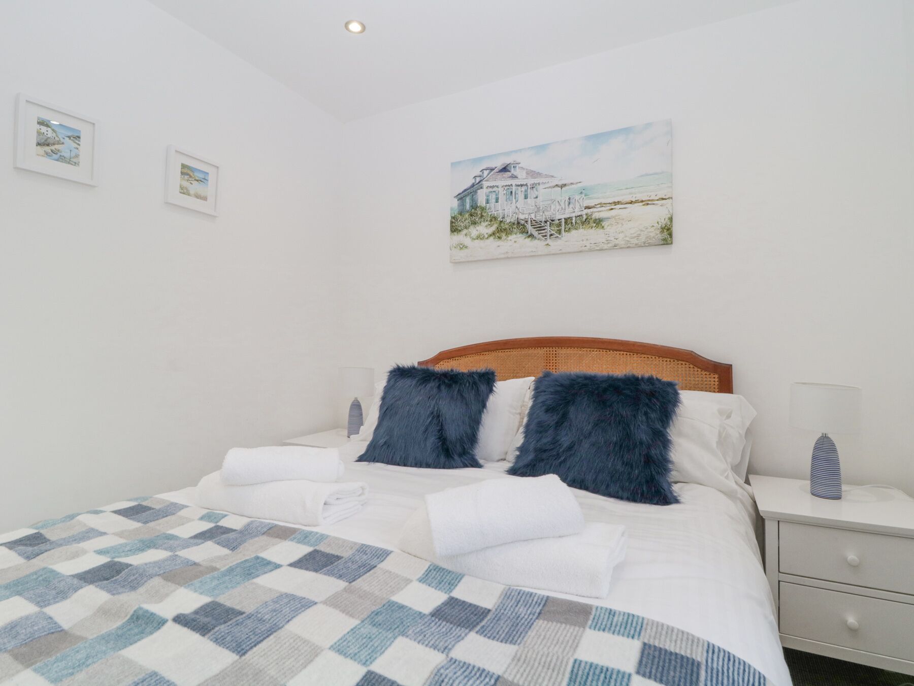 Una cama bien hecha con una colcha a cuadros, un cabecero de madera y dos almohadas azul oscuro. En la pared, encima de la cama, hay un cuadro de una casa de playa.