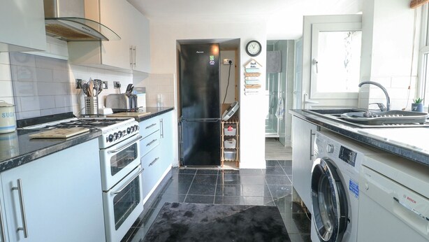 A modern kitchen with a stove, oven, refrigerator, and washing machine. There is a black rug on the floor, and a clock on the wall. - 63 Kingsland Road (Holyhead)