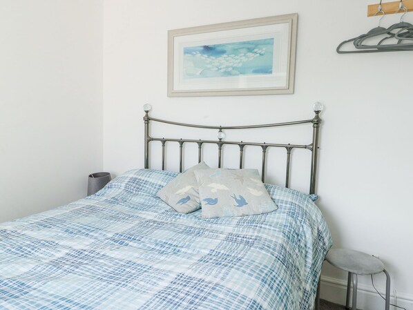 A bed with a metal frame and a blue checkered bedspread, a pillow with a bird pattern, a framed picture on the wall, and a coat rack with hangers. - 63 Kingsland Road (Holyhead)