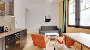 Un espace de vie compact comprenant un coin cuisine, un canapé gris, deux fauteuils orange, un miroir rond et une grande fenêtre avec des rideaux.