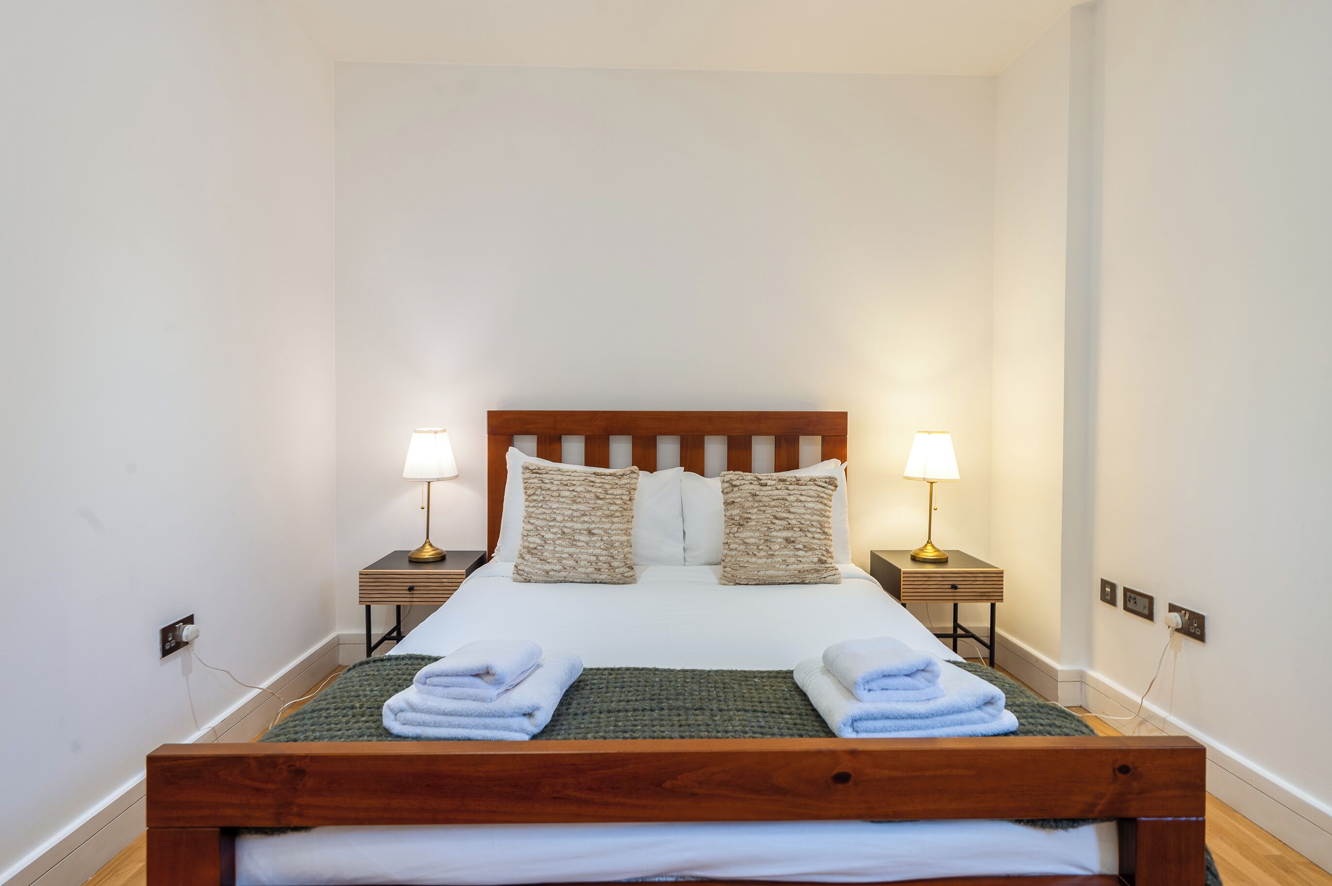 A neatly made bed with white linens and two pillows, flanked by bedside tables with lamps, in a simple room with white walls and wooden flooring.