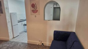 Un salon avec un canapé bleu foncé, une cuisine à l’arrière-plan et un petit miroir fixé au mur.