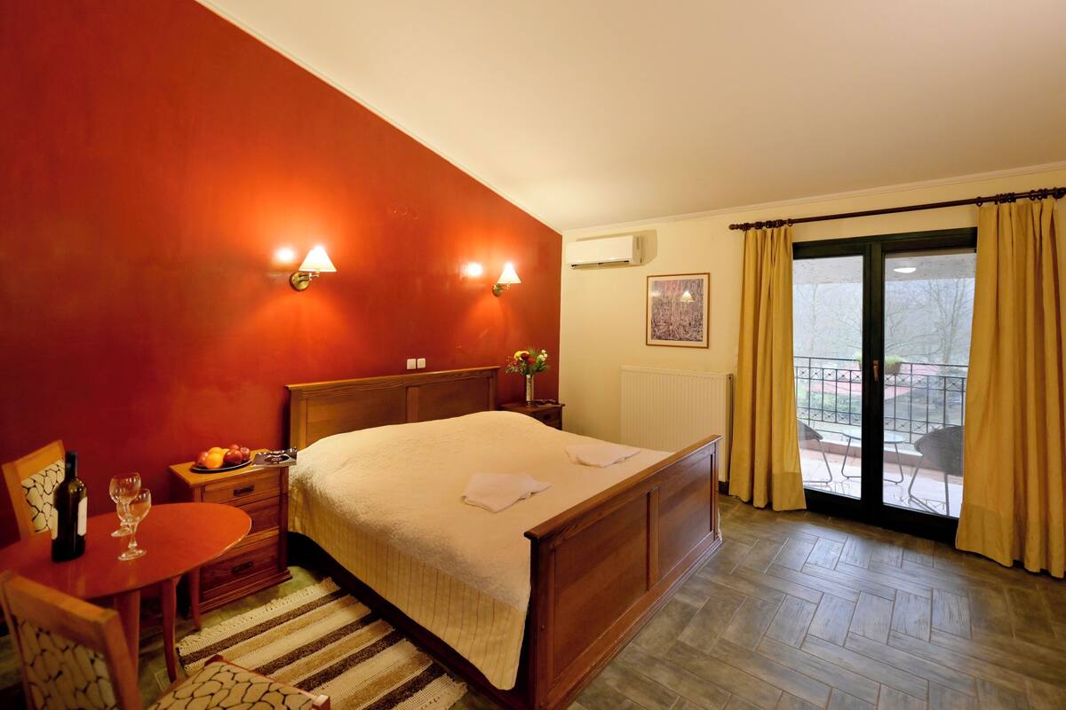 A hotel room with a bed, a small table with a bottle and glass, a chair, a window with curtains, and a view of the outside.