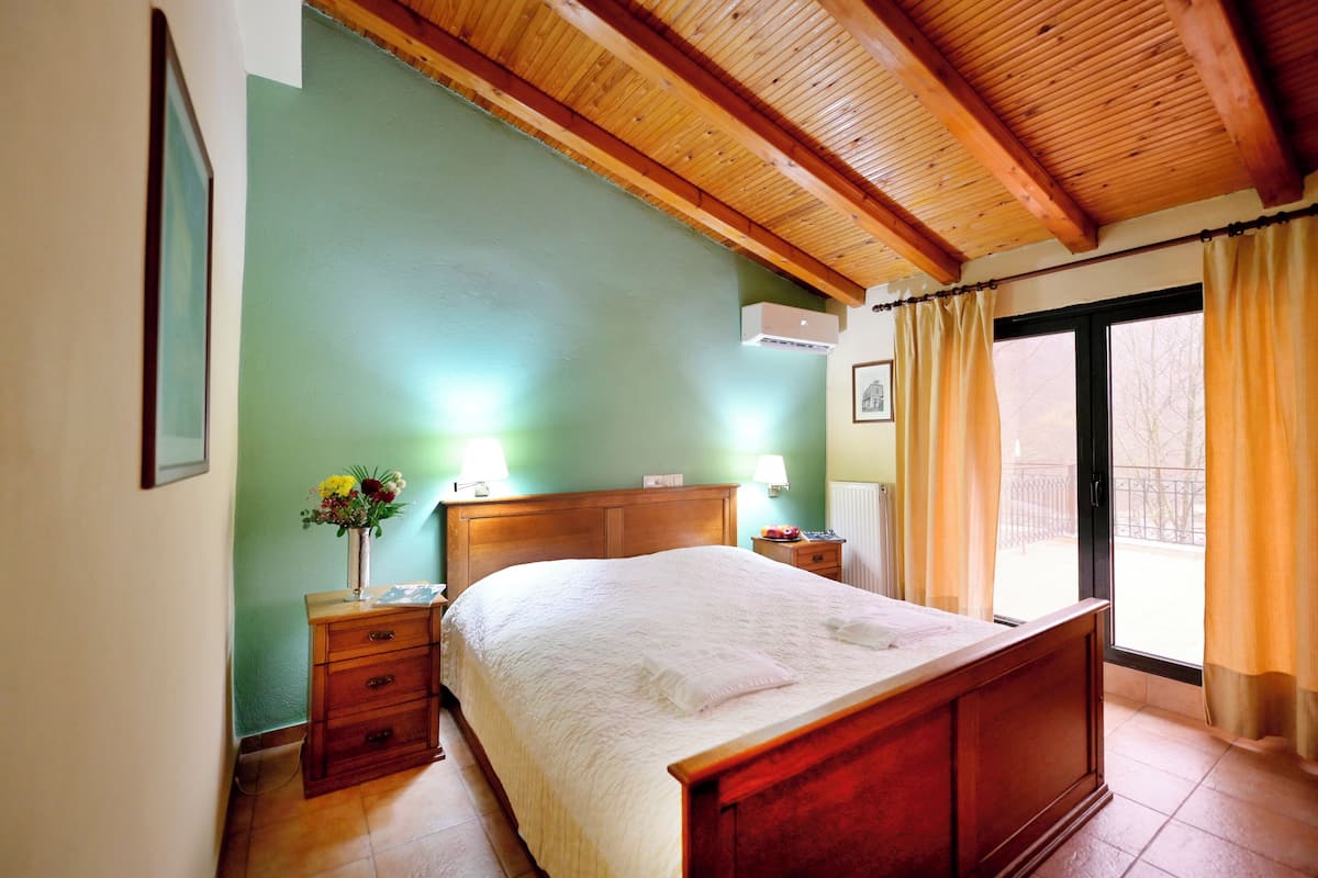 A bedroom with a wooden bed, a nightstand, a bedside table, a wall-mounted air conditioner, and a window with curtains.