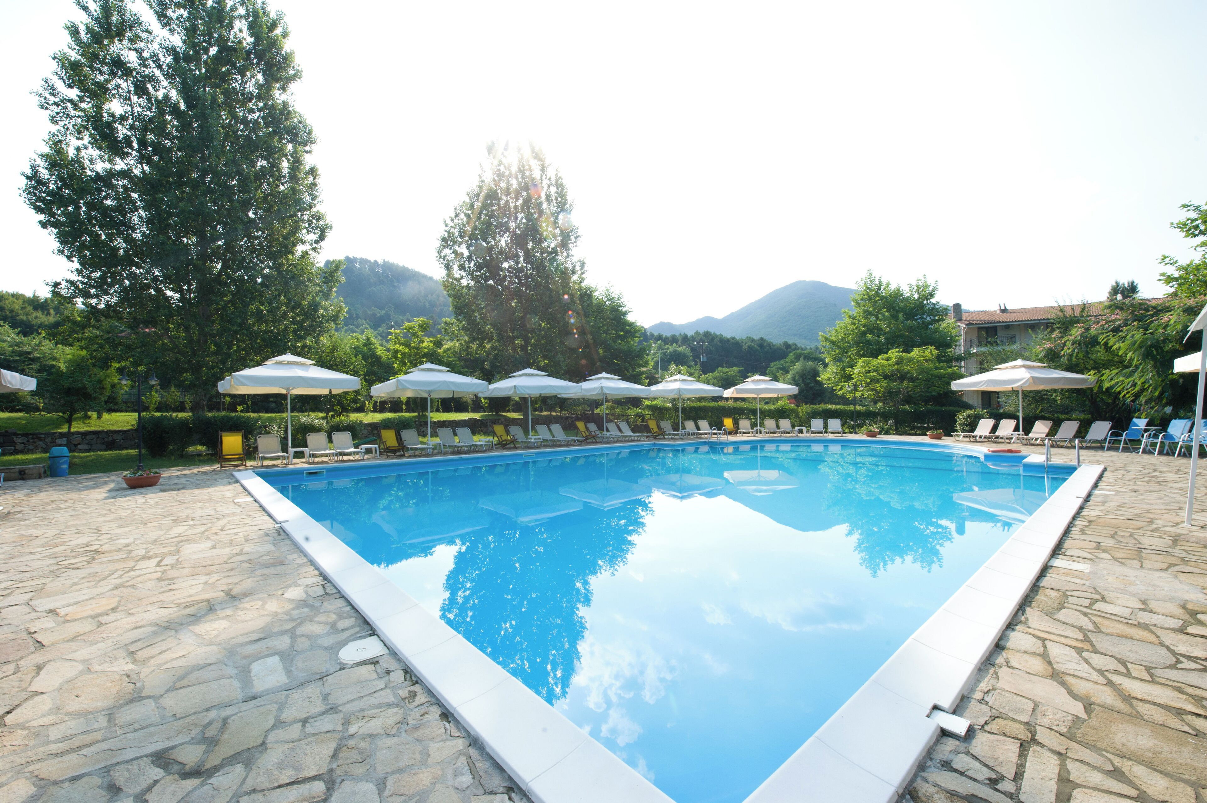 Une piscine entourée de chaises longues et de parasols, avec un sol en pierre et de la verdure tout autour, et des montagnes en toile de fond.