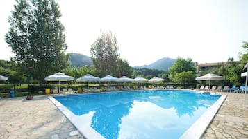 Une piscine entourée de chaises longues et de parasols, avec un sol en pierre et de la verdure tout autour, et des montagnes en toile de fond.