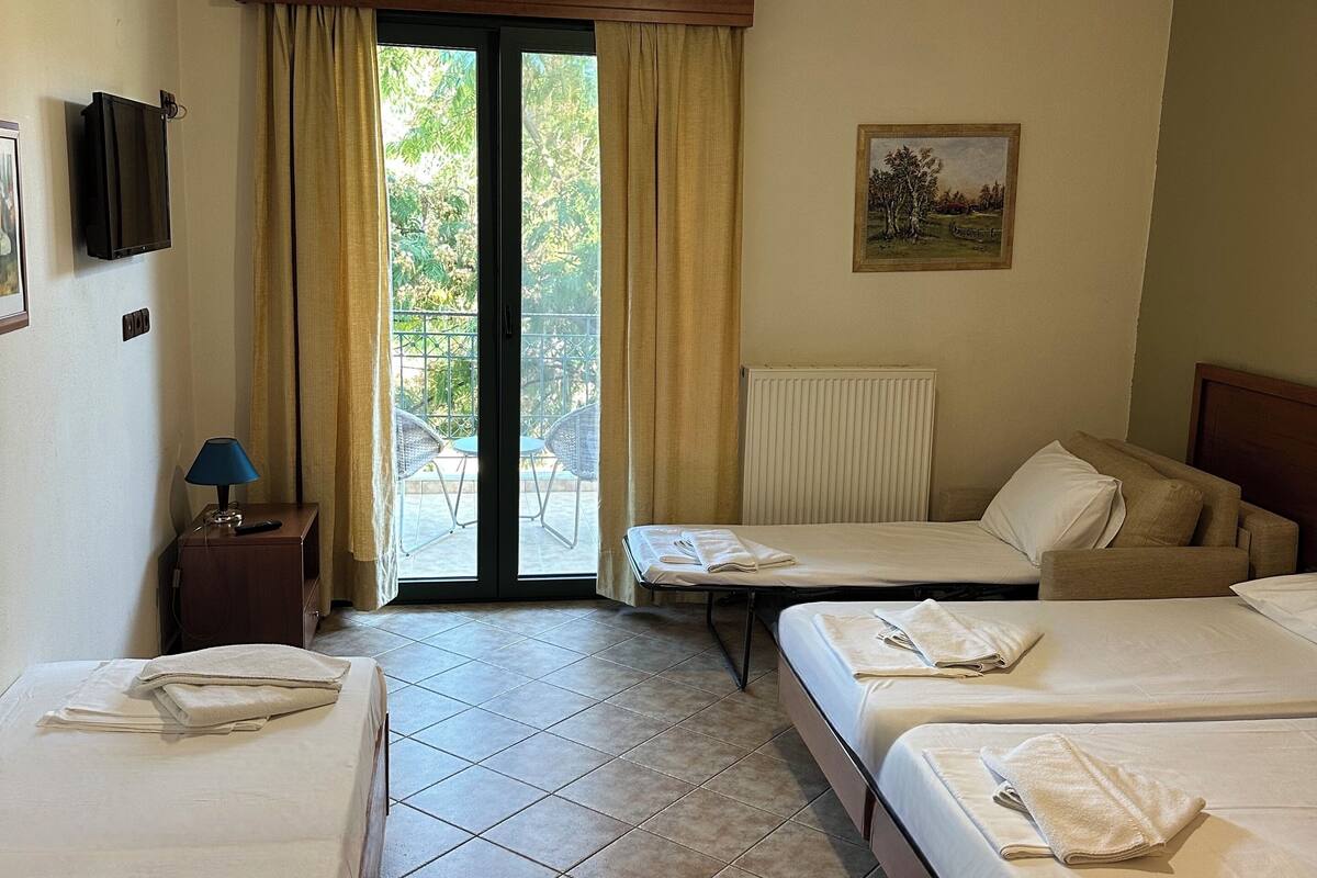 A hotel room with three beds, a ceiling fan, a painting, and a view of the outdoors through a sliding door.
