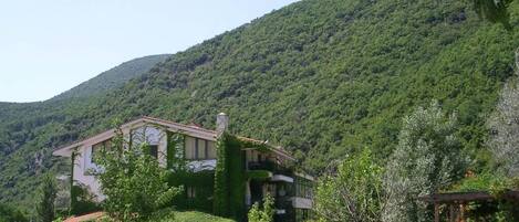A two-story building with a garden in front, surrounded by lush greenery and hills.