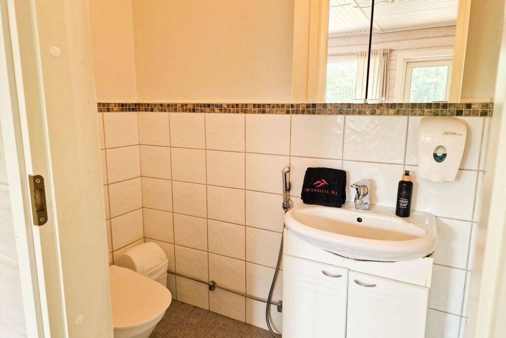 Une petite salle de bain propre, avec des carreaux blancs, un lavabo blanc et des toilettes. On y trouve un distributeur de savon et une pancarte noire accrochée au mur.
