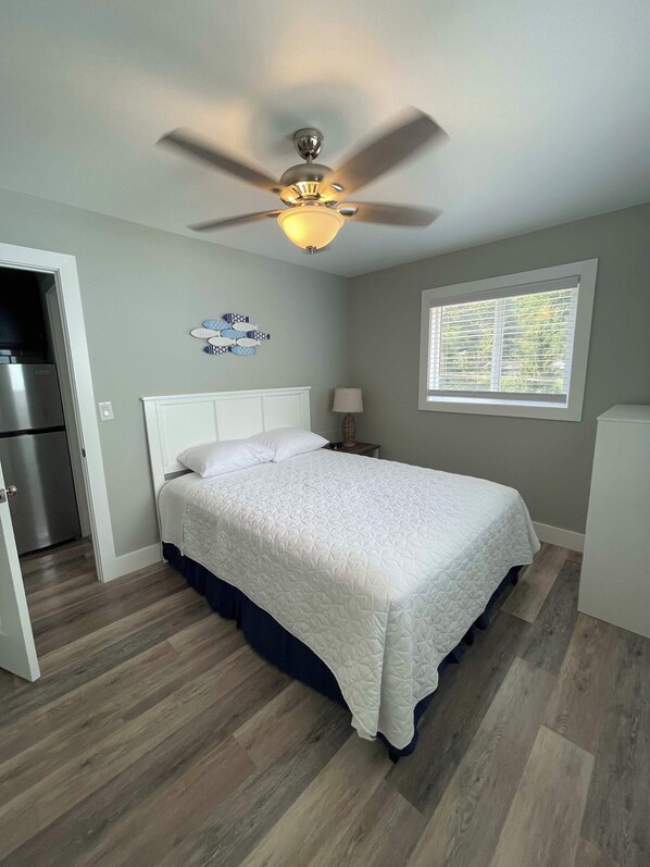 Un dormitorio con cama, ventilador de techo, ventana con persianas y refrigerador.