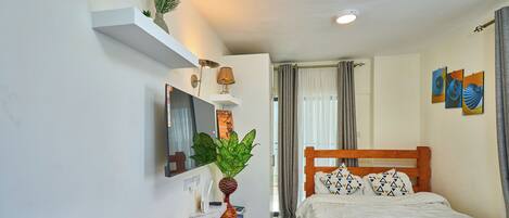 A hotel room with a bed, a desk with a computer, a mirror, a plant, and artwork on the wall.