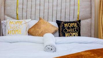 Una cama bien hecha con una manta marrón, dos almohadas con el texto 'HOME Sweet HOME' y una lámpara de pared.