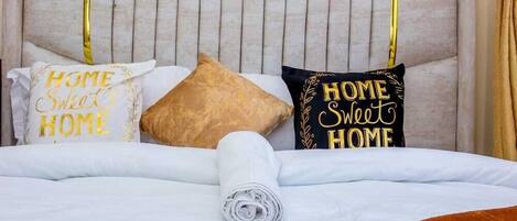 A neatly made bed with a brown throw blanket, two pillows with 'HOME Sweet HOME' text, and a wall-mounted light fixture.