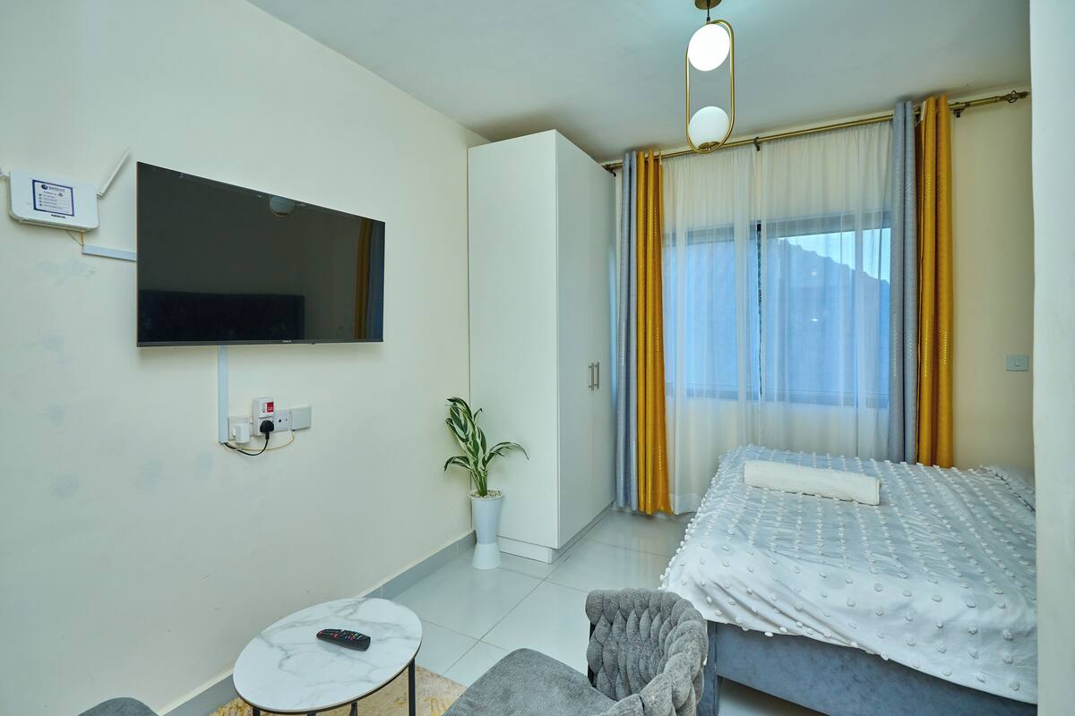 A modern hotel room with a flat-screen TV mounted on the wall, a small round table with a remote control, a grey armchair, and a bed with a white duvet.