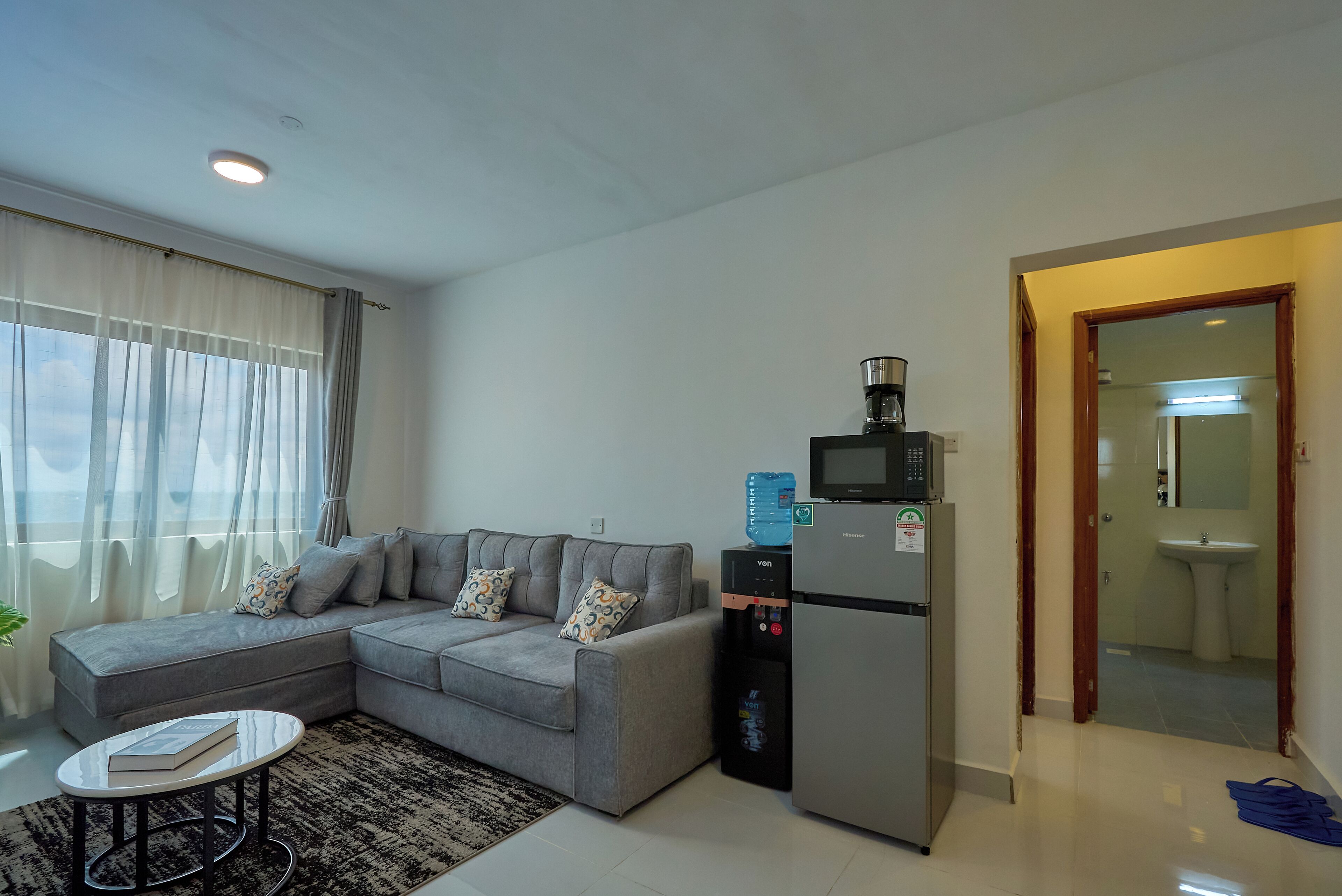 A modern living room with a grey sofa, a small round table, a refrigerator, and a water dispenser.