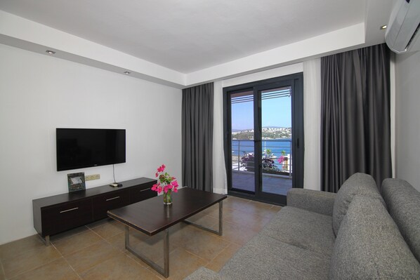 A modern living room with a flat-screen TV, dark wooden furniture, a large window with curtains, and a view of a cityscape. - Milport Hotel Bodrum (Bitez)