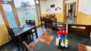 A dining area with wooden tables and chairs, a ceiling fan, and a view of a building outside. - Hotel Sakura Palace (Manali)