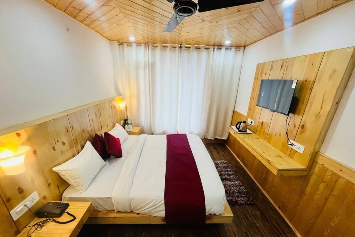 A bedroom with a wooden ceiling, a bed with white and red bedding, a bedside table with a lamp, a TV mounted on the wall, and a wooden bench.