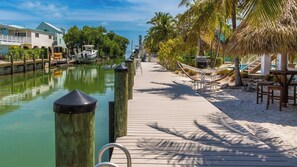 A wooden boardwalk leading to a beach with palm trees, a boat dock, and a building with a green roof.