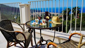 Un balcon avec des fauteuils en osier, une table en verre et une vue sur la mer et le paysage urbain.