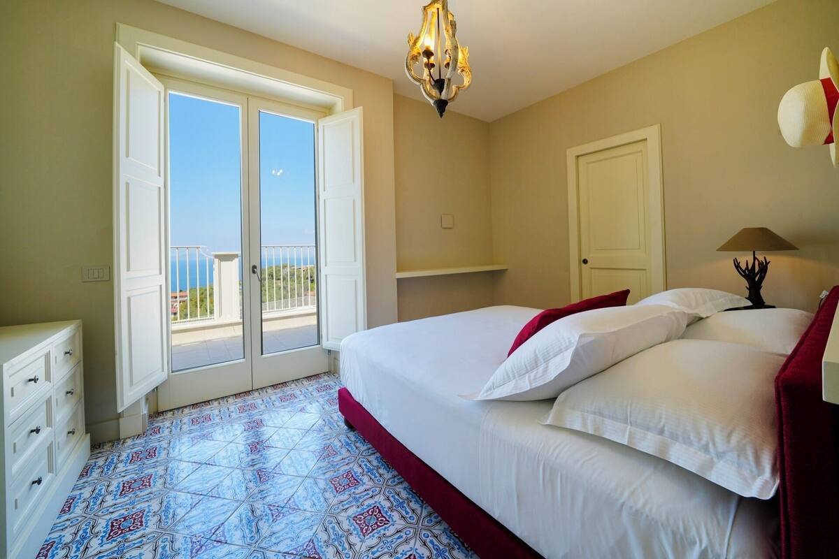 A bedroom with a large bed, a chandelier, a door, and a view of the sea through a sliding door.
