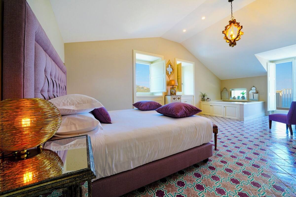 A bedroom with a large bed, a bedside table with a lamp, a dresser, and a view of the outside through a window.