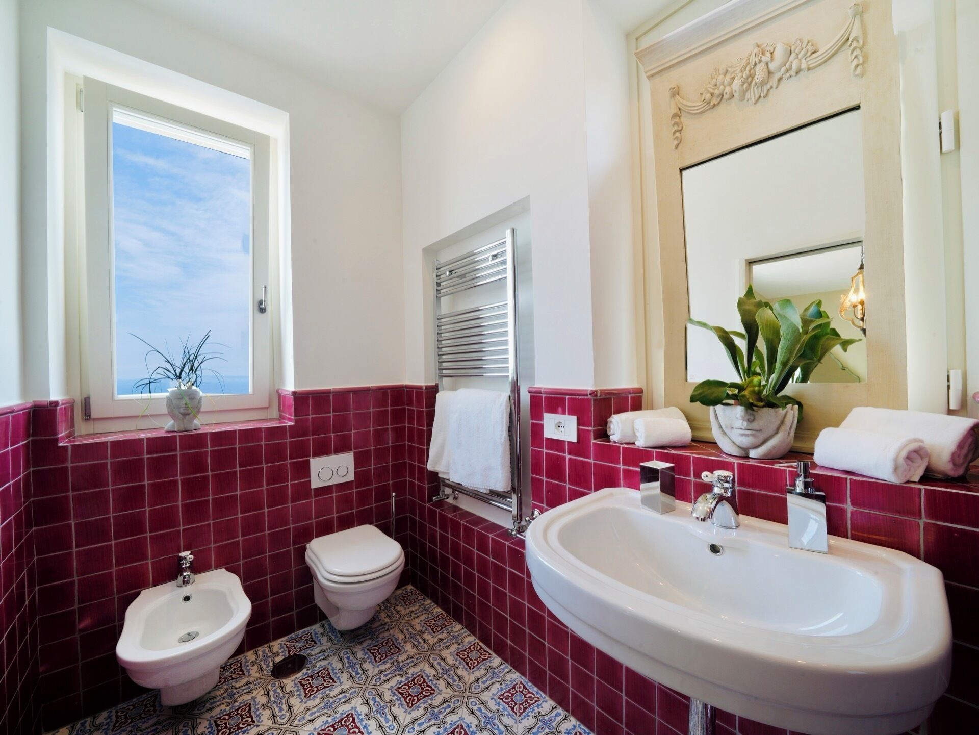 Une salle de bain équipée d’un bidet, de toilettes et d’un lavabo, avec des carreaux rouges et une fenêtre agrémentée d’une plante.