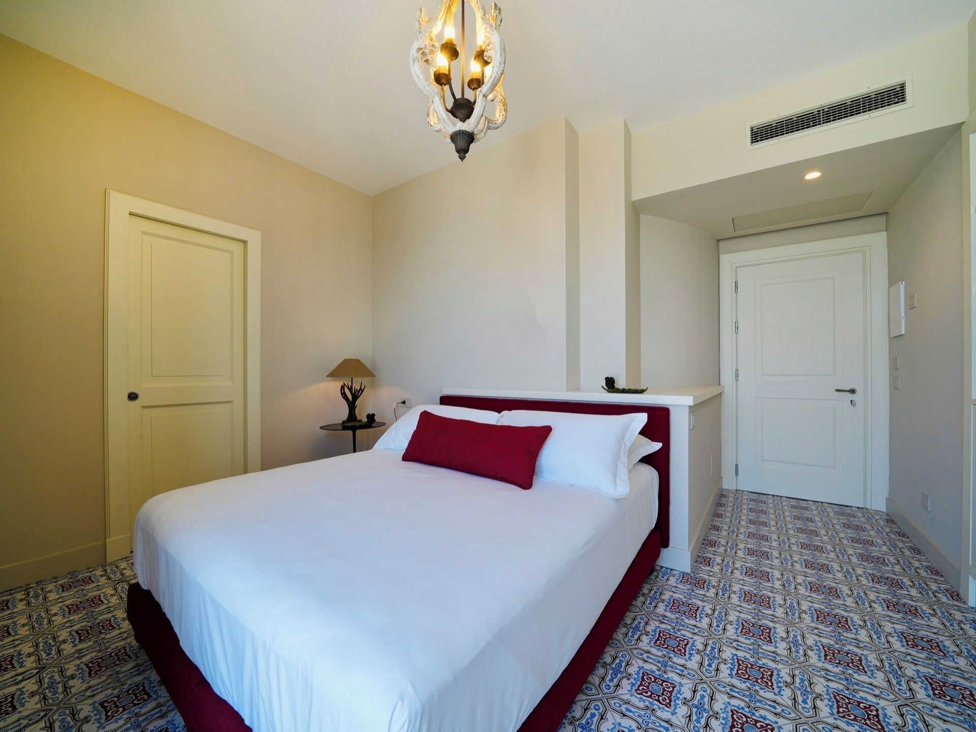 A hotel room with a large bed, a chandelier, a nightstand with a lamp, patterned carpet, and a door leading to another room.
