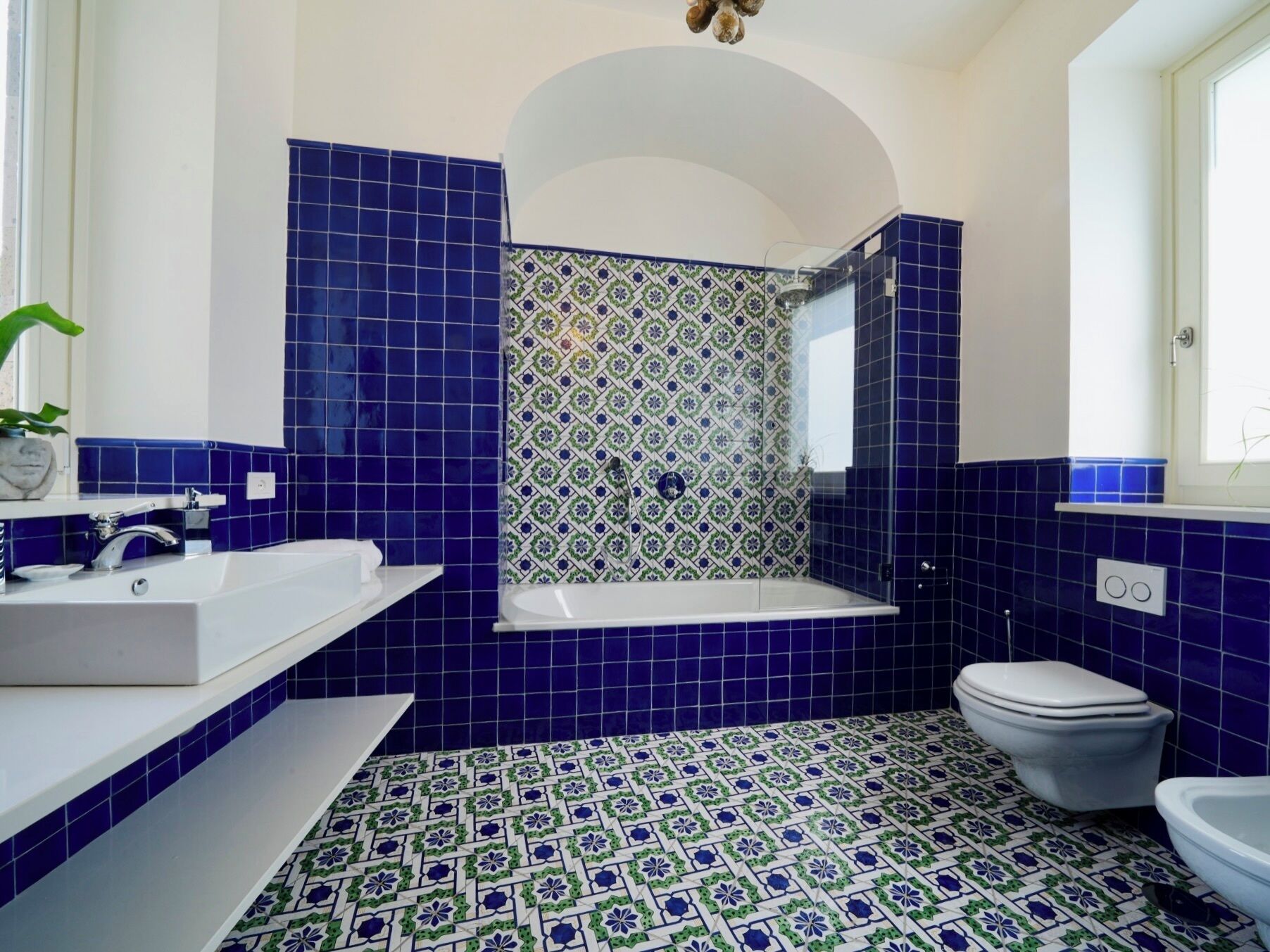 Um banheiro com pia branca, banheira e vaso sanitário. As paredes são revestidas com azulejos azuis, e o chão possui um design de ladrilhos estampados.