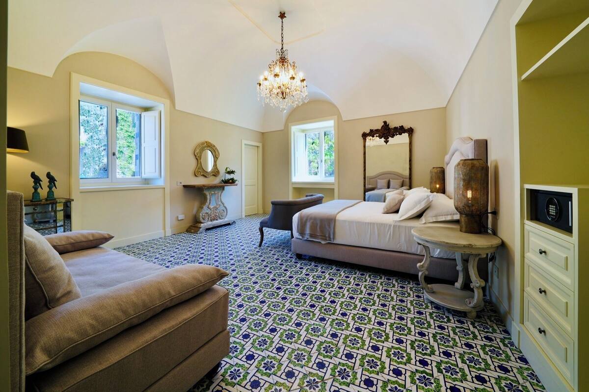 A bedroom with a large bed, a chandelier, a console table, and a window with a view of greenery.
