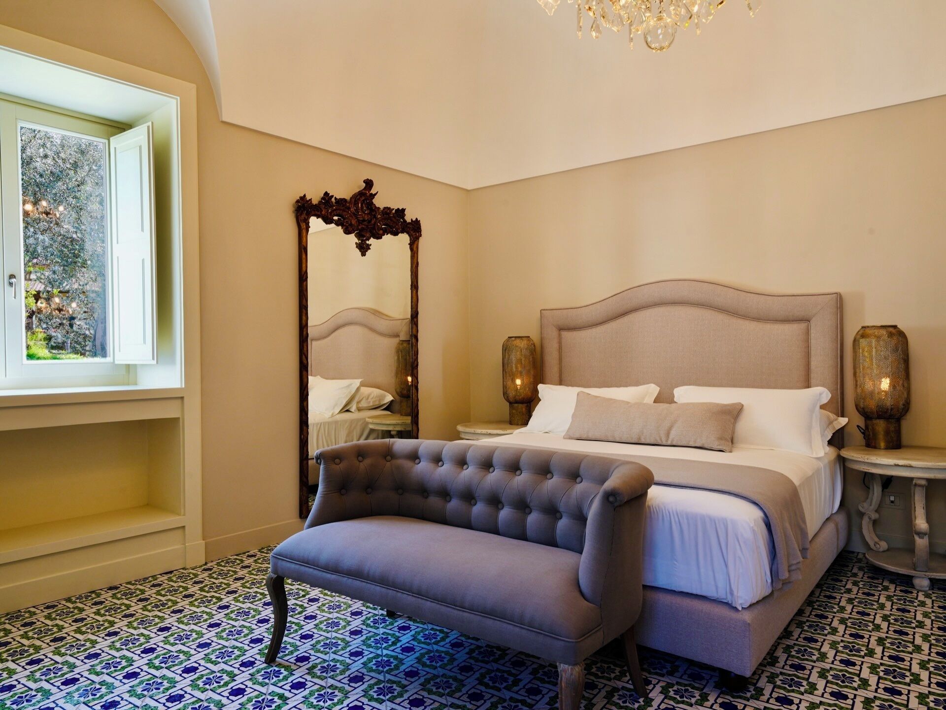 A bedroom with a large bed, a tufted armchair, a mirror, and a chandelier.