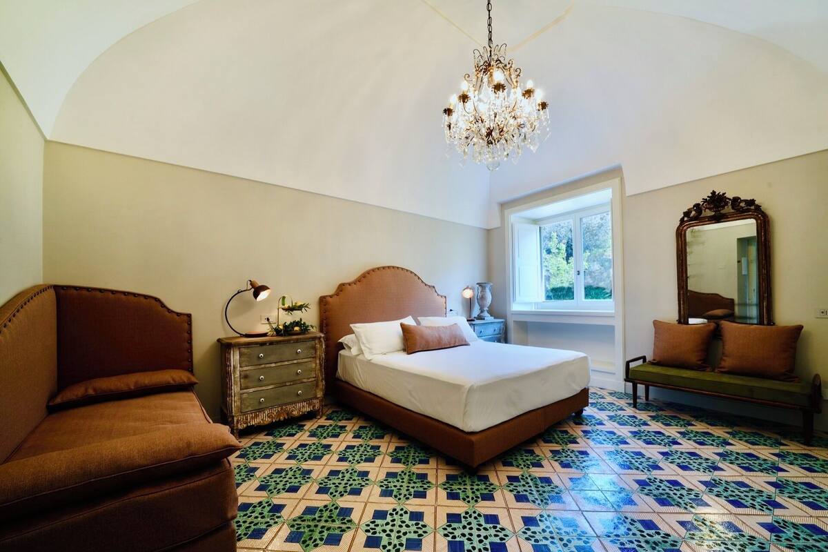 A bedroom with a large bed, a wooden dresser, a brown sofa, a chandelier, and a window with a view of greenery.