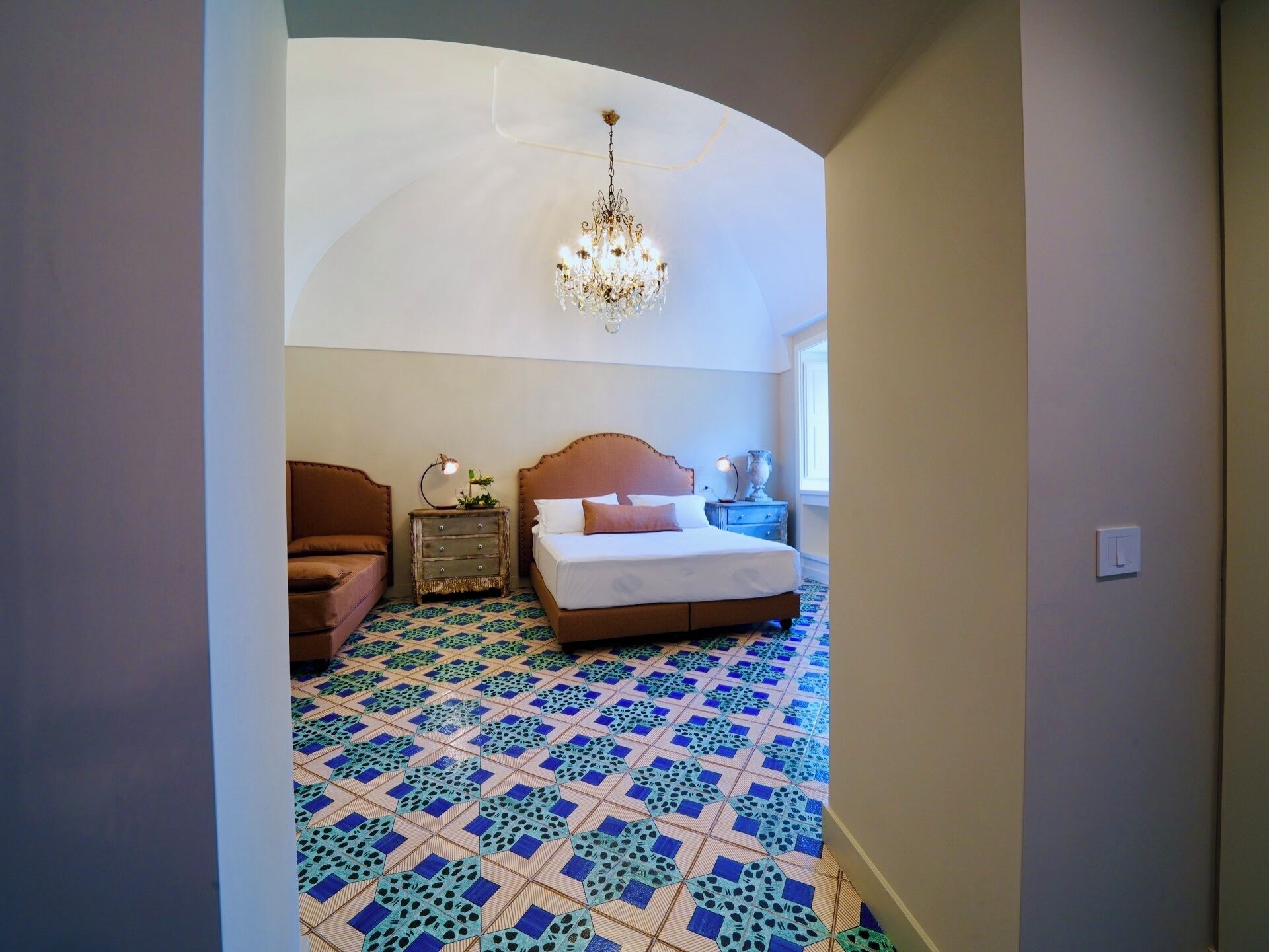 Camera da letto con un letto grande, un comodino, una sedia, un lampadario e piastrelle decorative al pavimento.