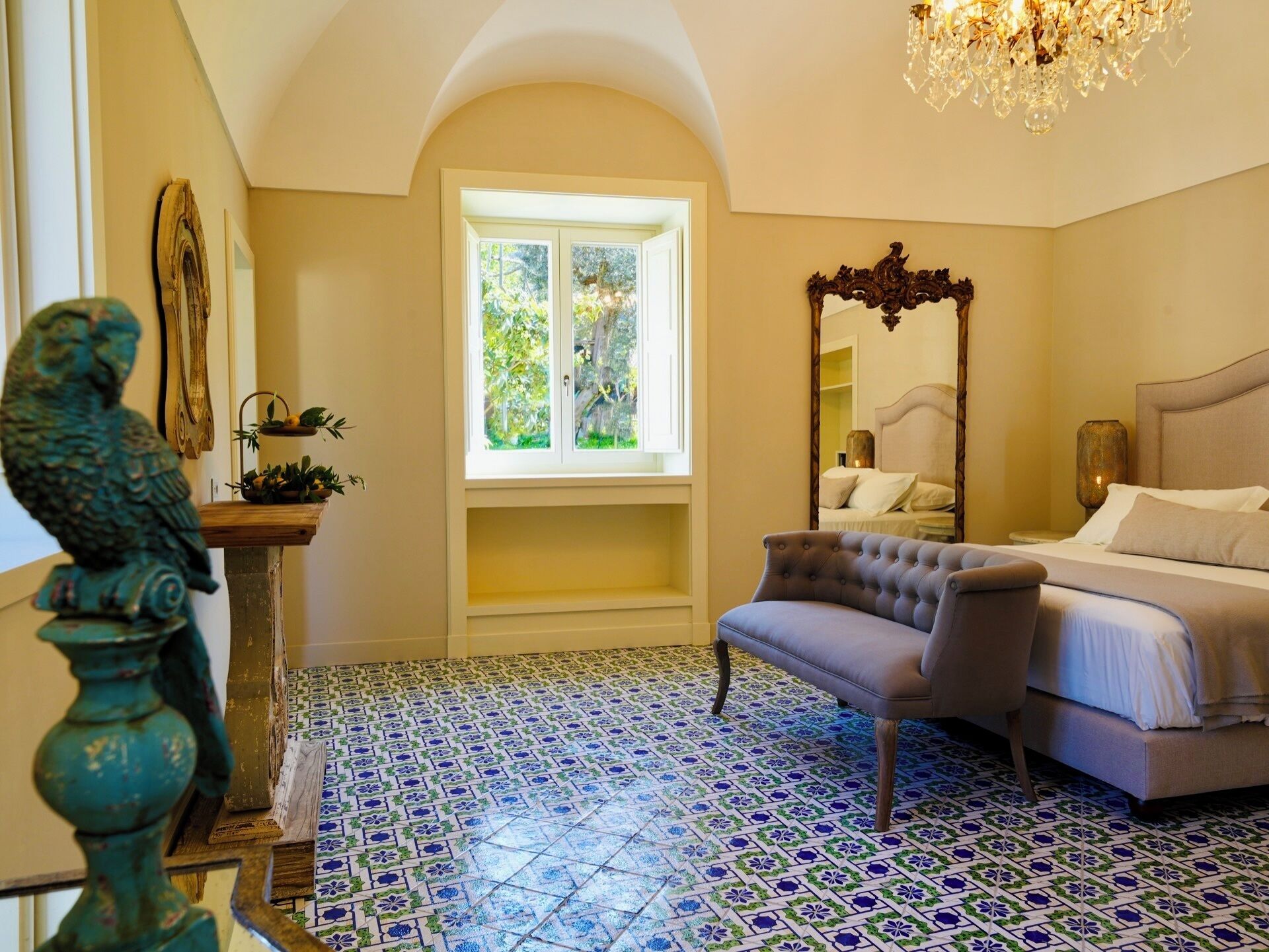A bedroom with a bed, a chair, a window, a mirror, and a chandelier.