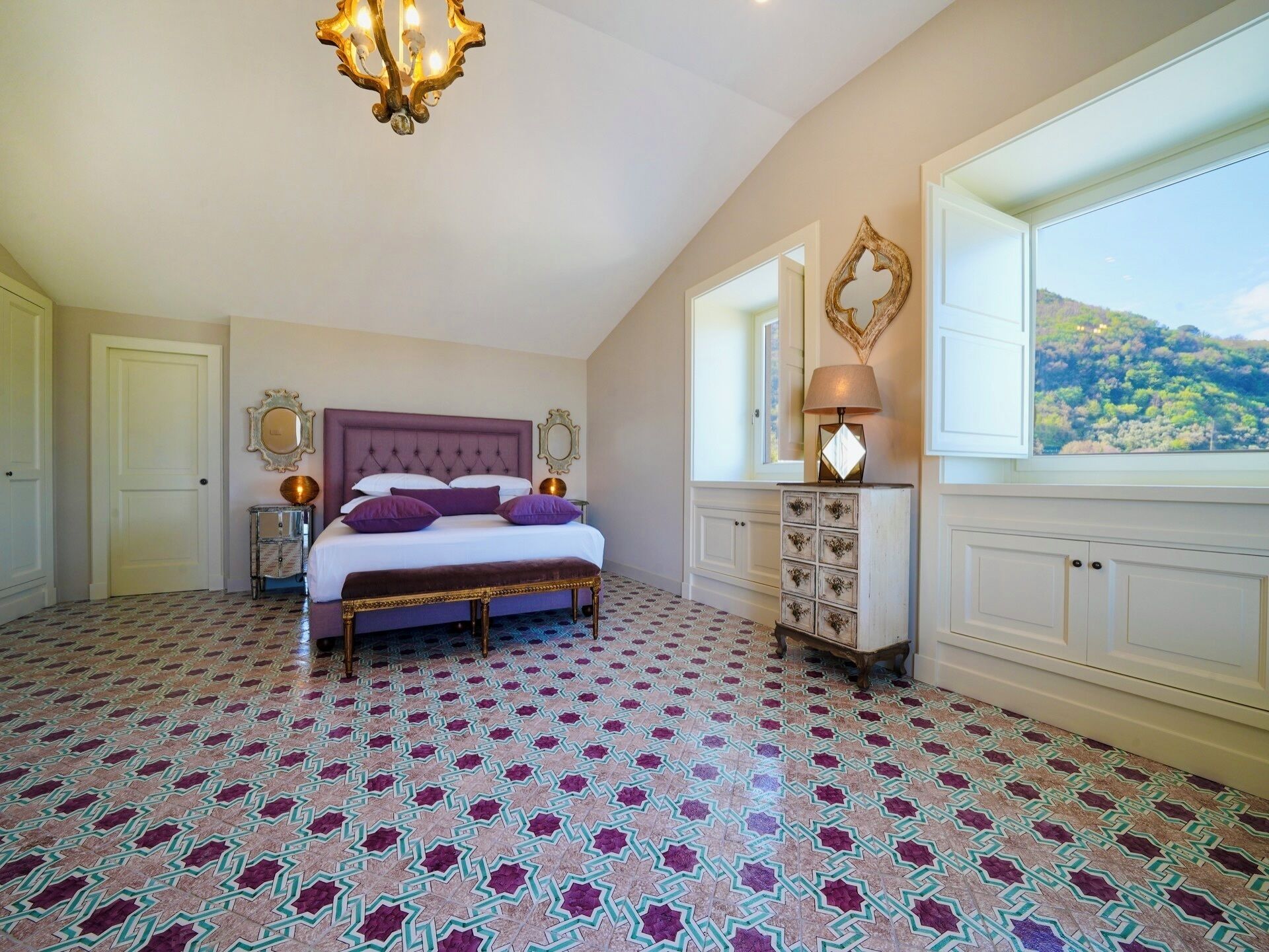 Une chambre à coucher avec un grand lit, un lustre, une table de chevet, une commode et une fenêtre donnant sur une colline.