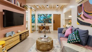 A modern living room with a flat-screen TV, a dining area with a table and chairs, and a sofa with cushions.