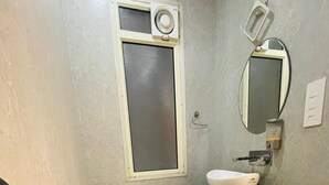 A bathroom with a toilet, a sink, a mirror, and a window with a ventilation unit. - Hotel Kapson Palace (Bareilly)
