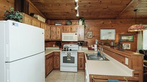 Une cuisine avec des meubles de rangement en bois, un réfrigérateur blanc, une cuisinière et un four à micro-ondes. On y trouve également un luminaire suspendu et une fenêtre avec des rideaux.