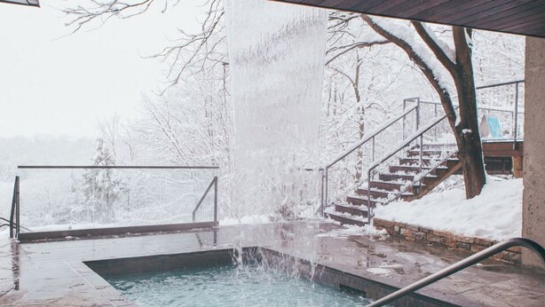 A hot tub with a waterfall feature, surrounded by a snowy landscape and a staircase leading to a covered area. - Charming Retreat in Eastman | Spa, Bike & Hike ! (Eastman)