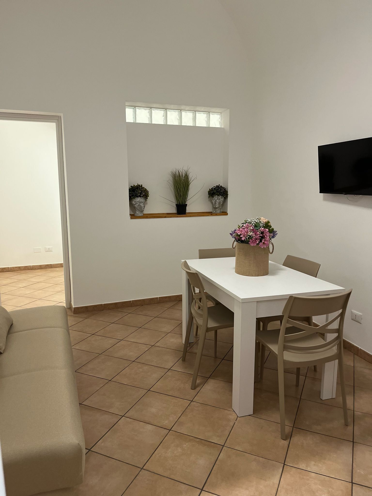 Et spiseområde med et hvidt bord og fire stole, en beige sofa, et fjernsyn monteret på væggen og et vindue med udsigt.