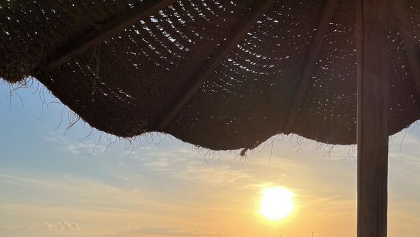 Une vue sur la plage avec un coucher de soleil, une structure dotée d’un toit de chaume et deux tabourets.