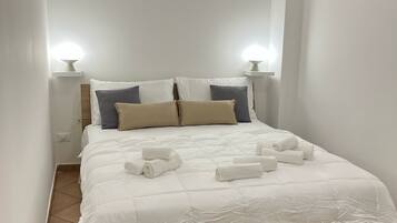 A neatly made bed with white bedding and rolled towels, a bedside table with a lamp, and a wall-mounted light fixture.